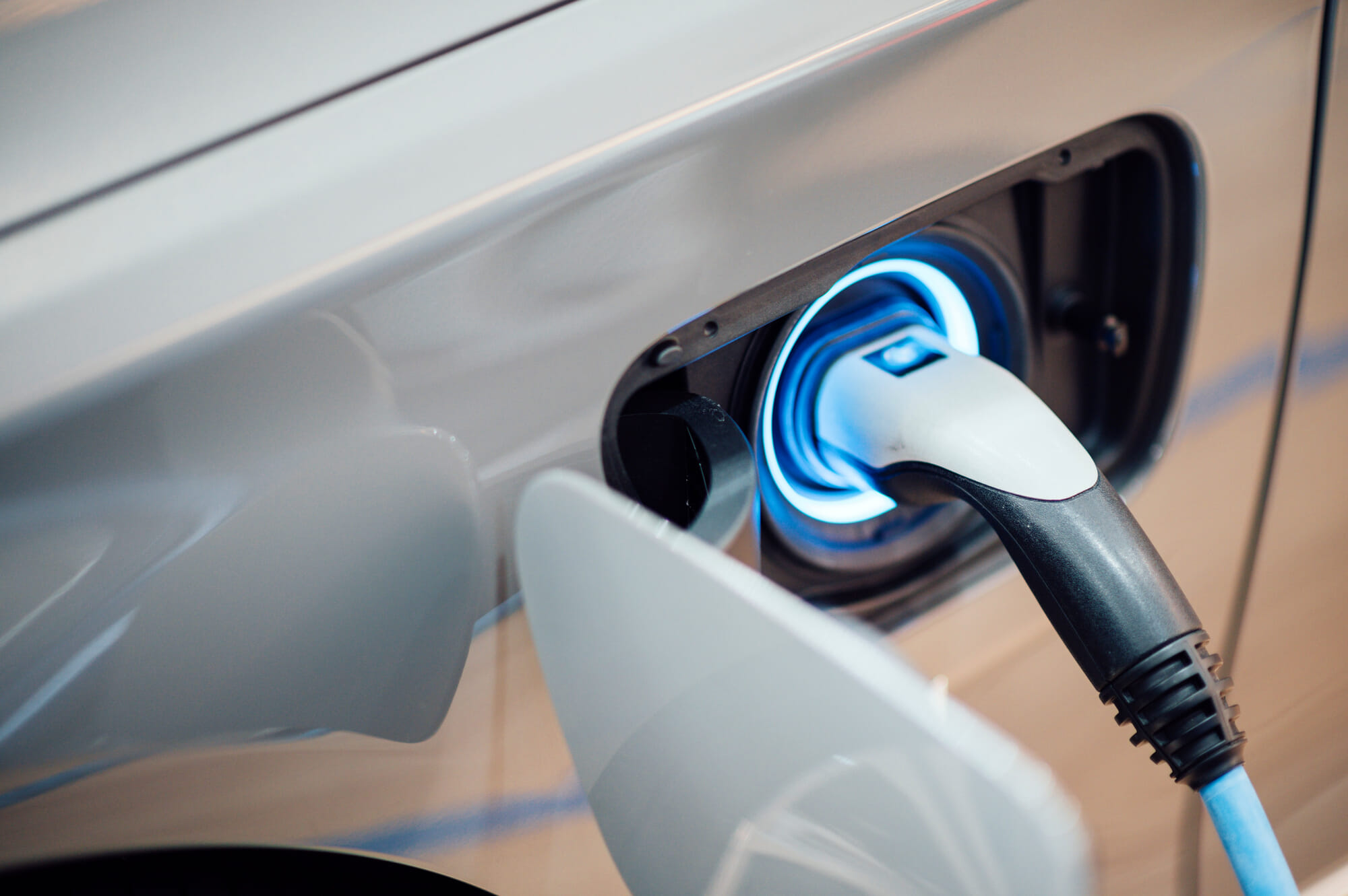 An electric charging station plug is connected to the charging port of a gray electric-powered car. The make and model of the car can't be determined from the photo.