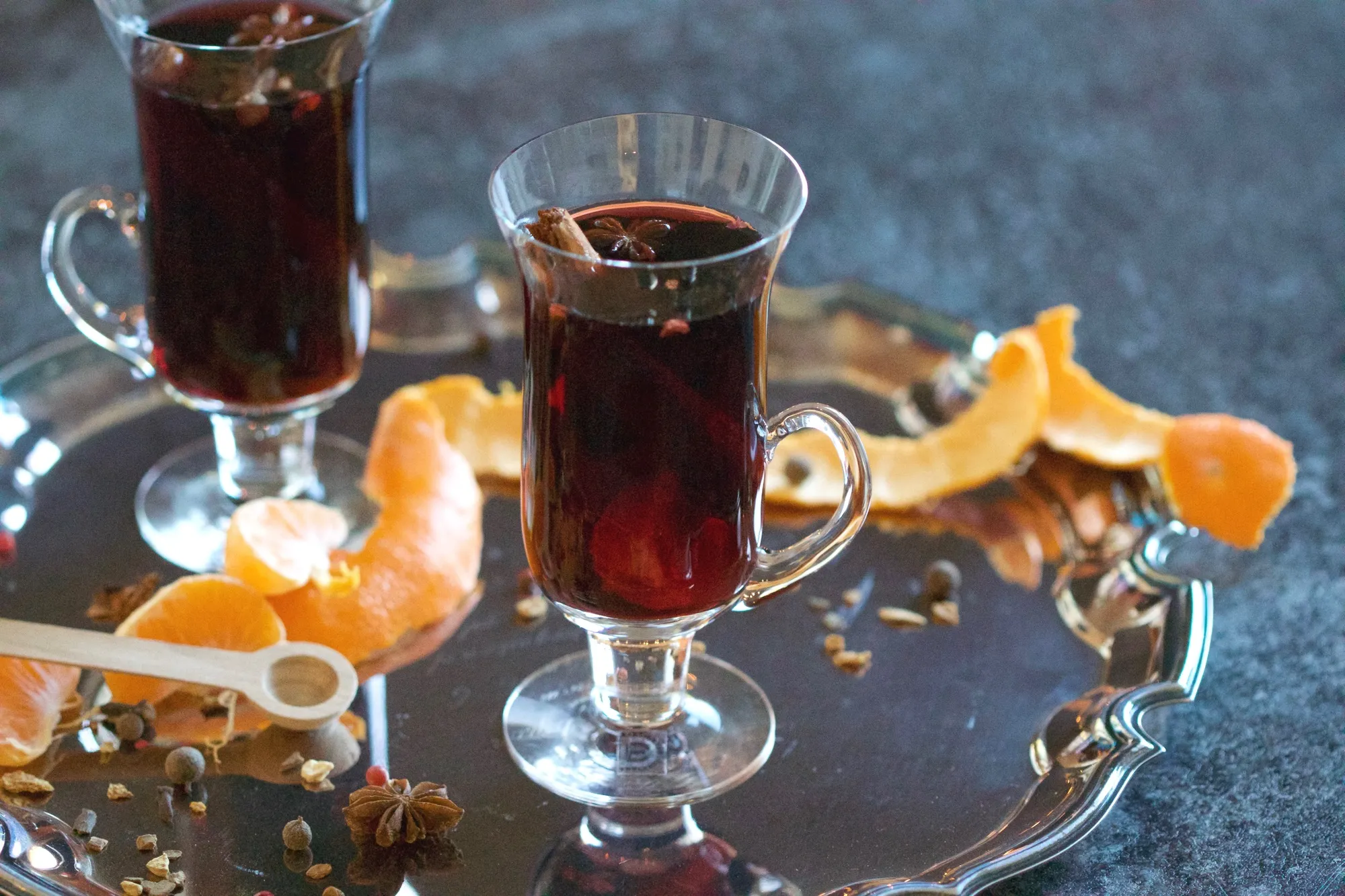 Glass mugs filled with mulled wine. The mugs are on a serving tray, and surrounded by orange peel and pieces of star anise and coriander seed.