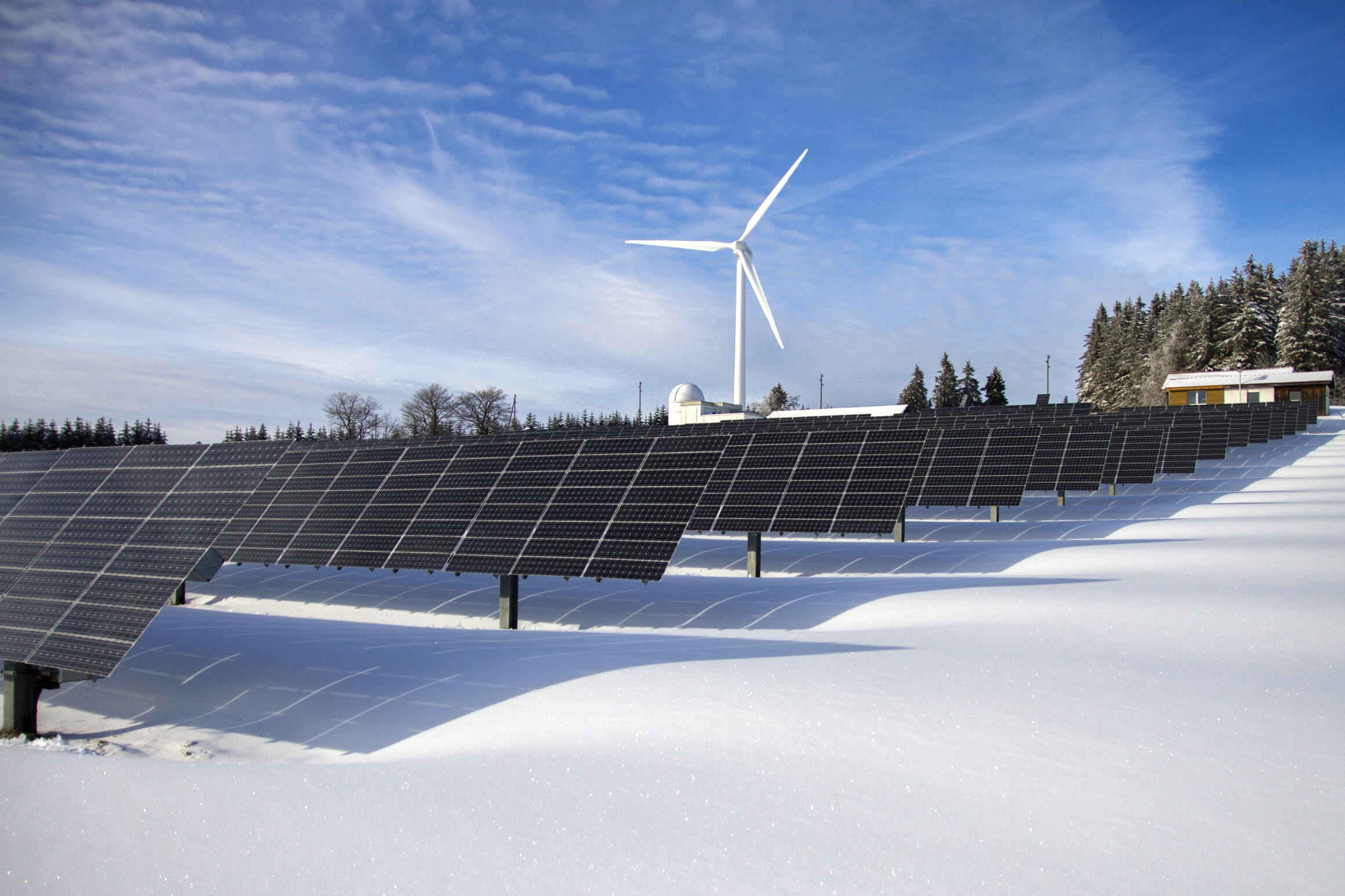 Solar panels in a snow-covered field with a wind turbine in the distance.