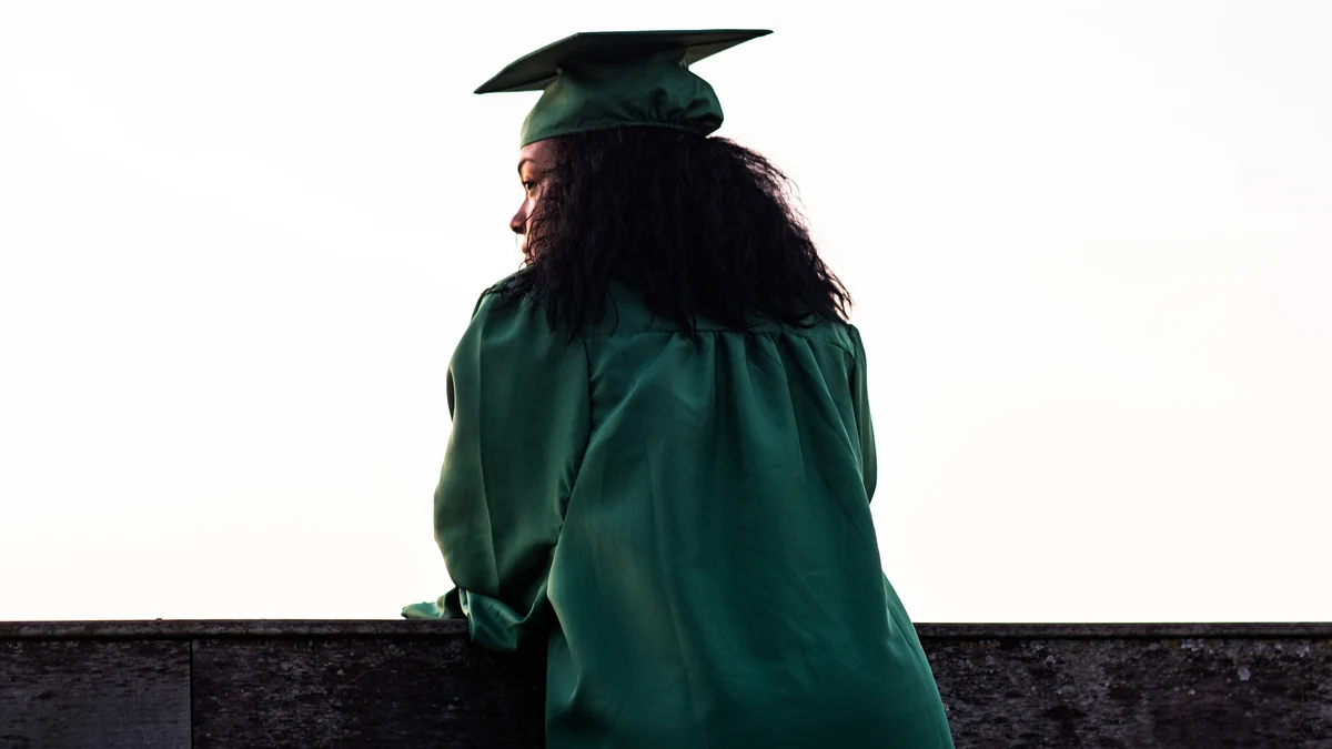 A woman wearing a hunter green mortarboard and matching robe leans forward on a wall, and looks into the distance. Her back faces the camera, but you can see her face in profile. She's standing against a bright gray sky.