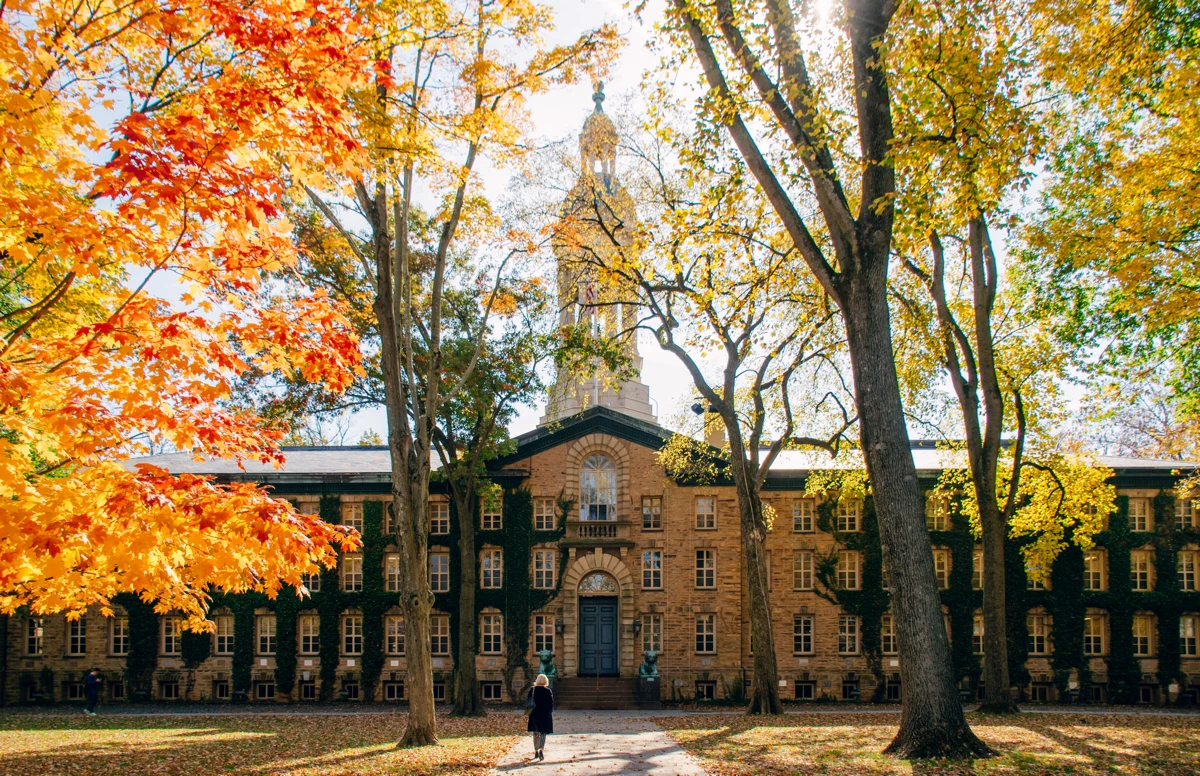 An old, yellowed brick building covered in ivy. A paved path leads from the foreground of the photo to the building's door. There's also a maple tree in the foreground of the photo. Its leaves are have turned orange, as they do in the transition to autumn. The grass on either side of the path is covered in fallen orange leaves from the maple tree and other trees. A feminine-appearing student walks the path.