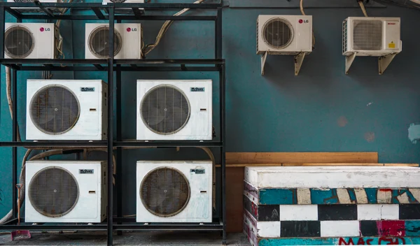 A rack of small air conditioners sits next to two air conditioning units that are perched on shelves. The wall behind the rack and shelves is painted teal.