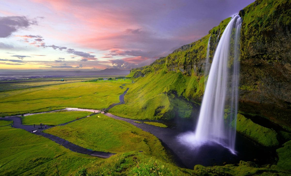 A medium-sized waterfall cascades over the side of a cliff. The cliffside is covered in a lush medium green carpet of moss and short grass. It's set against a sky at either sunrise or sunset. The clouds and angle of the sun means that the sky is streaked with light purple, orange, and yellow.