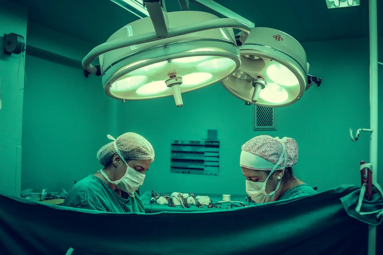 Two medical personnel wearing masks are performing tasks in an operating room. It's not clear whether this is an active surgery, pre-operative prep, or post-operative tasks.