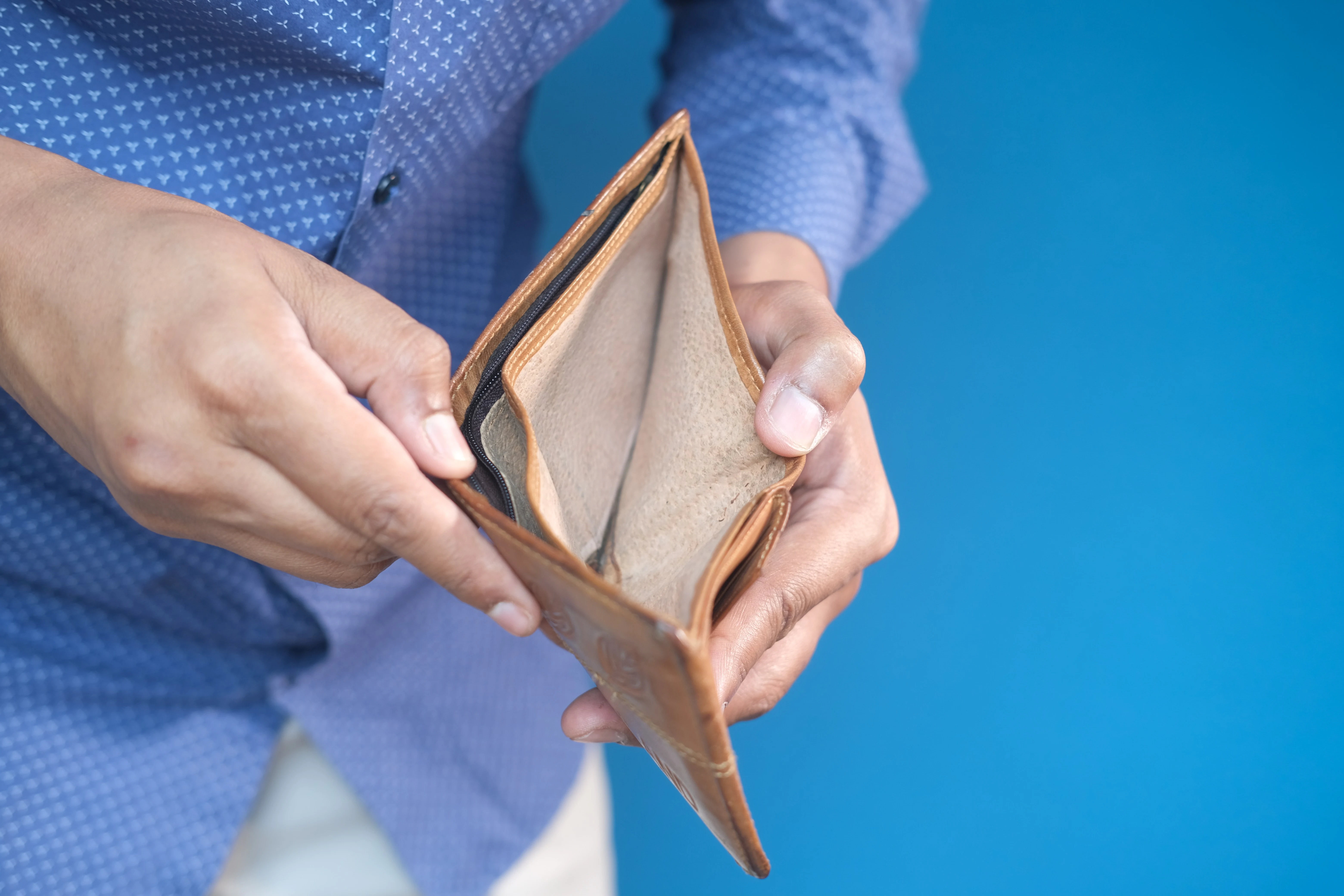 Closeup of two hands holding open a brown leather wallet. There is no money in the wallet. The person holding the wallet is wearing a blue button down shirt and cream colored pants. They are standing in front of a blue wall. Their face is not in the photo.