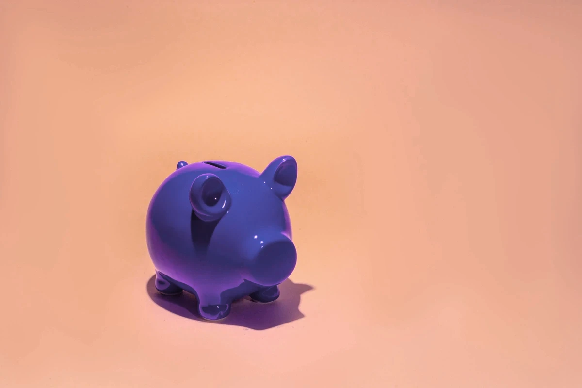 A small royal purple, ceramic pig-shaped bank set against a light peach or cantaloupe-colored background.