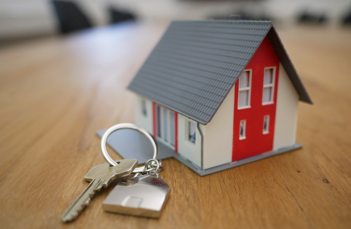 A miniature red and white house with a steeply-pitched roof sits on top of a wooden table. In front of the house there's a single key on a key ring. The key ring is attached to a house-shaped fob by a very short chain.