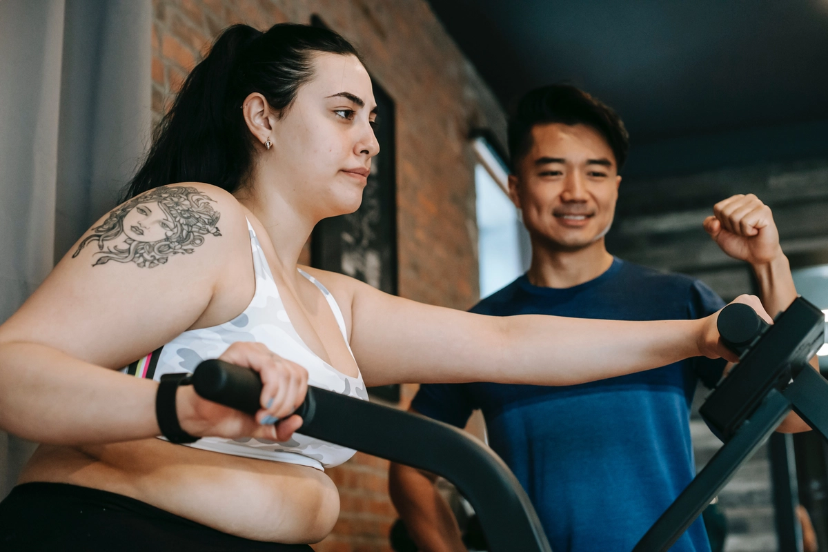 An overweight woman exercises on either an elliptical machine or an assault bike, and looks miserable while doing it. Her trainer tries to be encouraging.