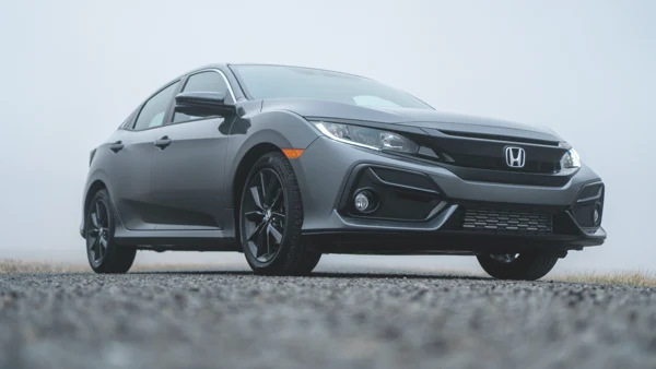 A late-model gunmetal-gray four-door Honda compact car is parked on asphalt near grass. The sky above it is gray with clouds. In the foreground of the photo, the asphalt is blurred.