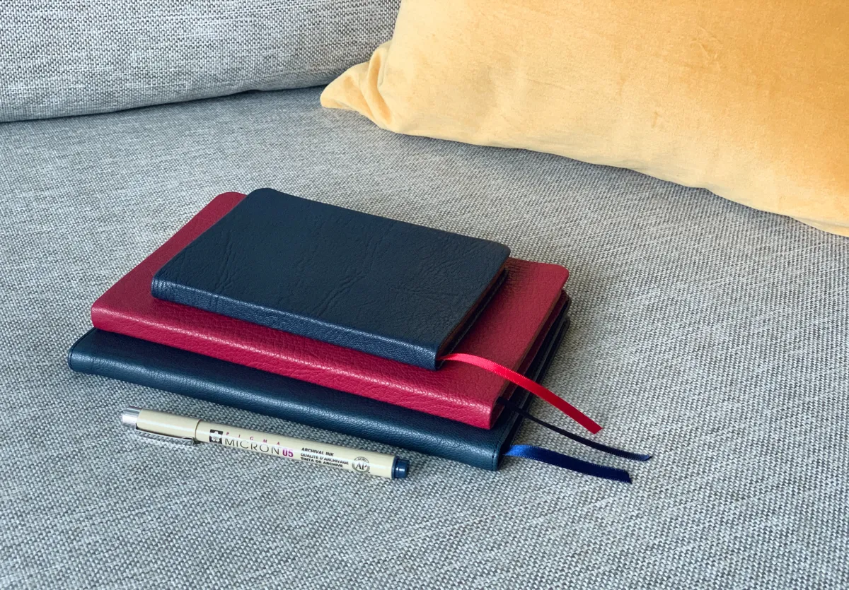 A stack of three leatherbound journals, two black and one red, stacked on a gray sofa, ready for writing.
